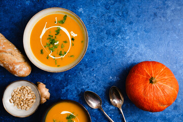 Pumpkin cream soup on blue background decorated with pumpkin, pumpkin seeds and fresh bread. Top overlay view. Autumn comfort food concept.