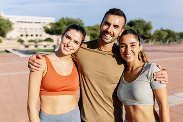 Portrait of three sportive friends smiling at camera embraced in an urban park in a sunny day