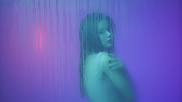 Portrait of female in neon light behind the glass window in steam and water drops. Girl with makeup and natural hairstyle posing hugging herself.