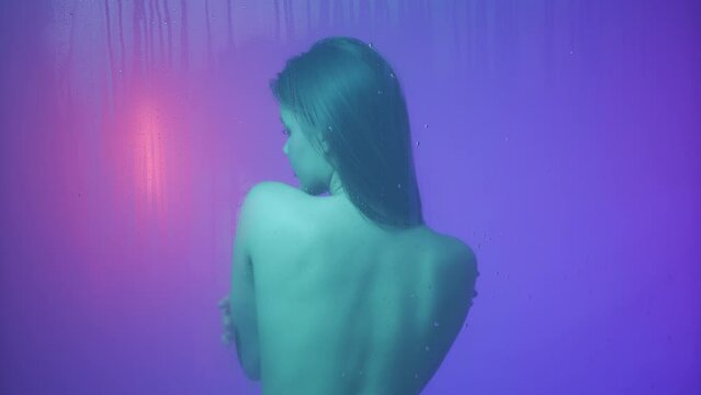 Portrait of female in neon light behind the glass window in steam and water drops. Girl with makeup and natural hairstyle showering behind glass.