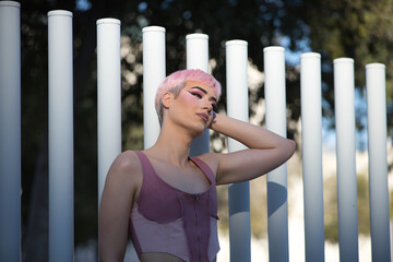 Young, attractive, gay man, heavily makeup, with pink hair and top, looking at infinity, leaning on white bars, receiving the last rays of the sun. Concept lgtbiq+, gay, pride, makeup, fashion, trend.