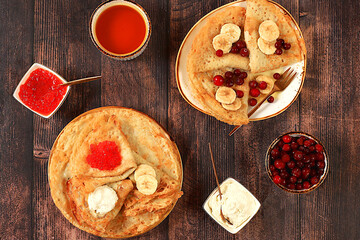 Delicious Russian pancakes with red caviar, soft cheese curd, bananas, cranberries and honey on a wooden table. Healthy breakfast with ingredients, carnival concept and traditional national cuisine,
