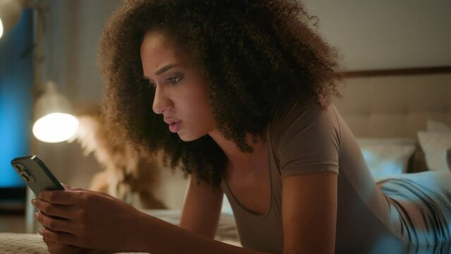Disappointed sad African American woman looking at mobile phone device upset frustrated ethnic female gen z girl lying on bed with smartphone worried scam message loss error failure at evening home