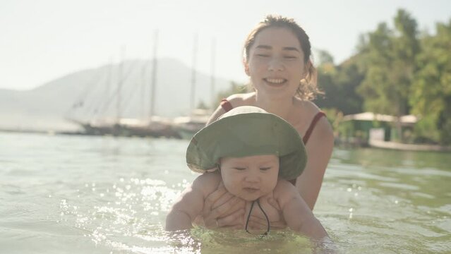 Mixed race family on vacation. Beautiful woman mother teach to swim her little baby boy, laugh and smile. Soft touch, love care, protection of mom and child relationship. Mom and cute baby rest by sea