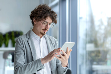 Businessman young thinking serious and concentrated standing near the window inside the office, the man is holding a tablet computer in hands, using application, passionately reading from screen.