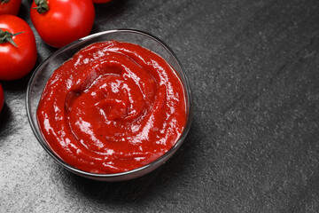 Organic ketchup in bowl and fresh tomatoes on black table, above view with space for text. Tomato sauce