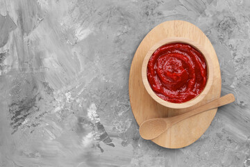 Organic ketchup in wooden bowl and spoon on grey textured table, top view with space for text. Tomato sauce