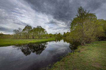 The river reflects the dramatic sky, a spring motif with the river and green bushes and grass.