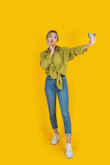 Sending air kiss, full body length young blonde caucasian beautiful woman sending air kiss. Taking selfie on mobile phone, smartphone. Isolated yellow studio background. People lifestyle concept idea.