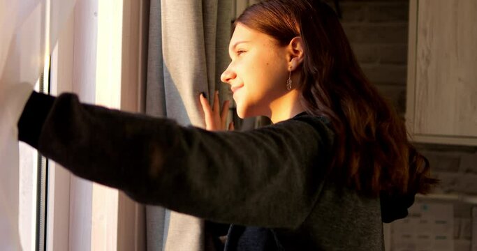 A young girl looks at the evening sun through the window. Open the curtains to see the rays of the winter sun.