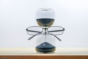 Keep an eye on time, hourglass with black sand wearing glasses, business concept of deadline running out, wooden table against a light gray background, copy space
