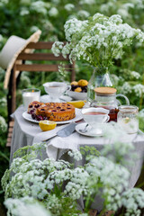 Tea time with sweet homemade cherry cake and herbal beverage in summer garden outdoors. Table covered with linen tablecloth, white porcelain cups. English style