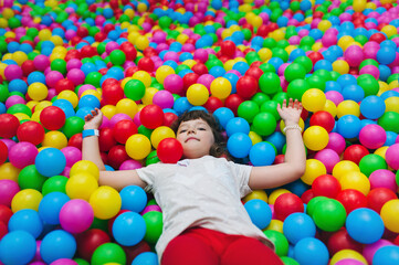 Fototapeta na wymiar A joyful cute curly girl in a white T-shirt and red pants lies in a pile of colorful bright balls in the game center and looks up.