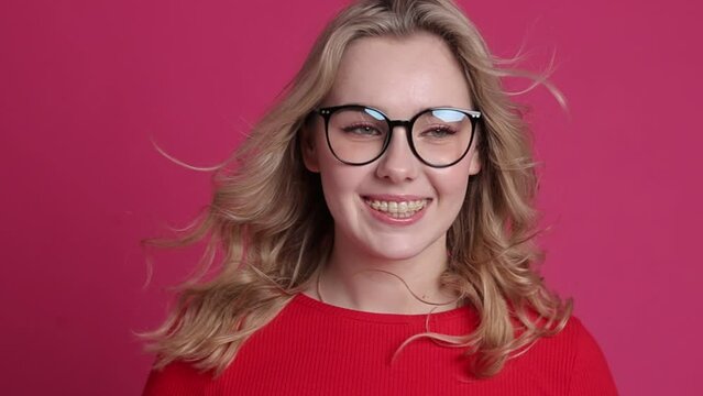 Cheerful young caucasian woman with transparent ceramic braces on teeth isolated on pink backdrop. Blond girl in eyeglasses looks at camera. Windy effect with fan. Beauty, health and fashion concept.