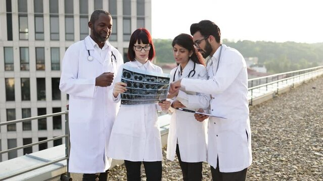 Doctors discussing x-ray image. Team of four professional male and female multiracial doctors examining patient's tomography scans while standing outside the modern hospital building.