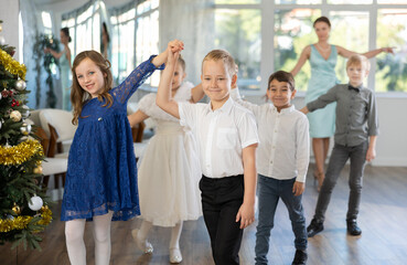Merry junior schoolchildren in party dresses lined up in pairs during dance in hall embellished...