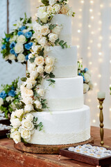 A four-tiered white wedding cake decorated with white roses on a wooden table and LED lights in the background