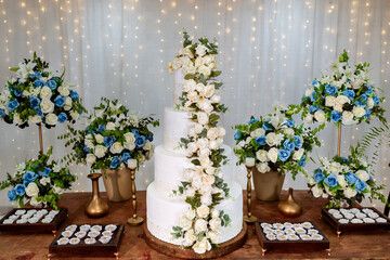 A four-tiered white wedding cake decorated with white roses on a wooden table and LED lights in the background