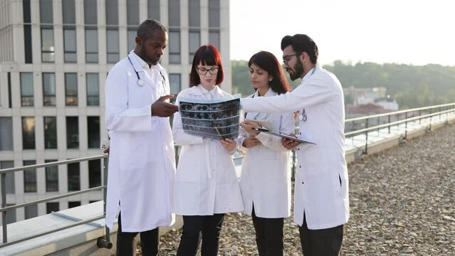 Team of four professional male and female multiracial doctors examining patient's tomography scans while standing outside the modern hospital building. Doctors discussing x-ray image.
