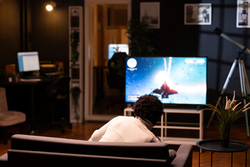 Man playing entertaining spaceship videogame on smart TV, relaxing on living room couch. Player...