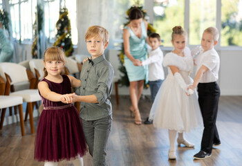 Happy little children in elegant dresses practicing waltz dance with teacher in school hall decorated with Christmas-tree