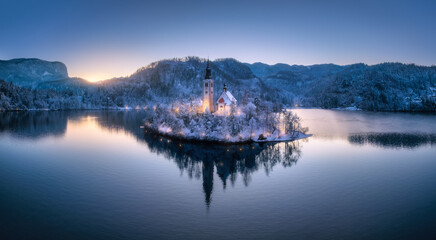 Aerial view of church on snowy island on the Bled Lake, Slovenia at winter night. Top drone view of chapel, alpine mountains, illumination, trees in snow, lights, reflection in water at sunset. Dusk
