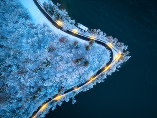 Aerial view of road, snowy forest, illumination, lake, street lights at winter night. Top drone...