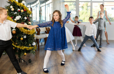 Enthusiastic tween girl in blue dress practicing partner jive moves in dance studio adorned with Christmas tree, under guidance of female teacher..