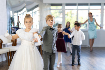 Fototapeta premium Group of cheerful emotional tweens, girls ad boys in formal wear dancing slow romantic partner dance in sunlit classroom during children festive event