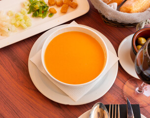 Cold soup gazpacho in bowl served with vegetables and baked breads on table in restaurant