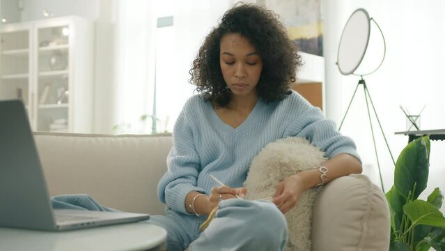 Curly female thinking through creative content plot writing. Concentrated multi ethnic girl writing down life or debt release plan. Focused woman on sofa. Elegant casual lady making notes in notebook