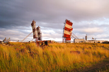 Dilapidated radar towers stand among tall grasses against a dramatic sunset sky, depicting a scene...