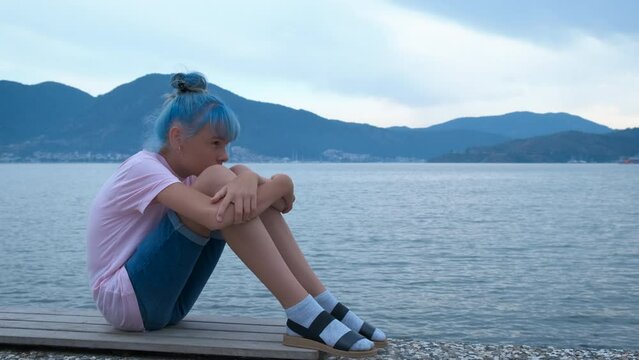 Loneliness of a teenager. A young girl with blue hair sits in a closed position on the shore of the sea bay.