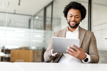 A content African-American businessman uses a tablet in the office, his smile reflecting the...