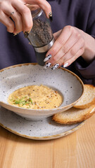 A woman pours pepper into a plate of cheese cream soup with nuts and greens.