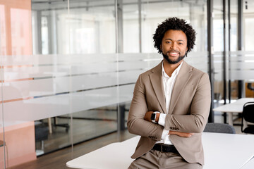 A confident professional in a taupe suit leans against a white table in a bright office setting,...