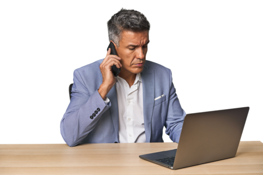 Serious middle-aged Hispanic man on phone while working on laptop