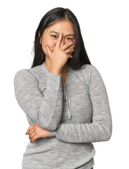 Young chinese woman isolated blink at the camera through fingers, embarrassed covering face.