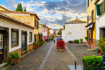 Tourists walk down the cobblestone Rua de Santa Maria narrow street of cafes, colorful doors and shops in the historic medieval old town of Funchal. - obrazy, fototapety, plakaty