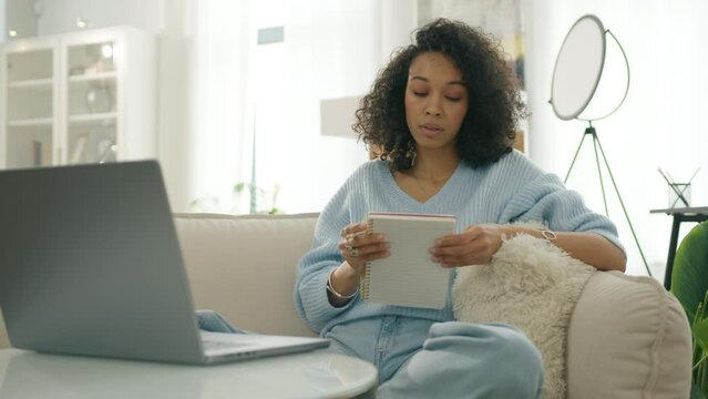 Elegant casual lady making notes in notebook. Curly female thinking through creative content plot writing. Concentrated multi ethnic girl writing down life or debt release plan. Focused woman on sofa