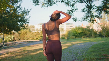 Fitness woman stretching neck walking summer back view. Lady warming up muscles