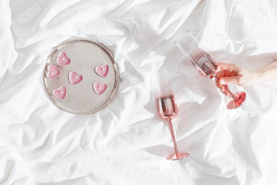 Top view Pink shining champagne glass in female hand, candles as hearts on bed cloth. Lifestyle aesthetic photo, star filter. Valentine's Day, love concept, romance mood. Sparkling wine.