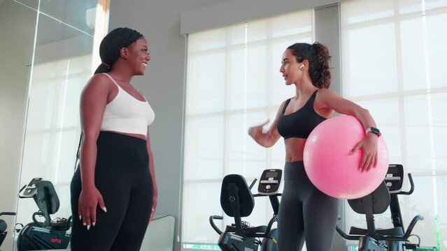 Two young woman in sportswear workout routines using exercise ball in fitness gym with trainer, instructor explaining and training exercise with woman for stretch muscles hand, sport and yoga.