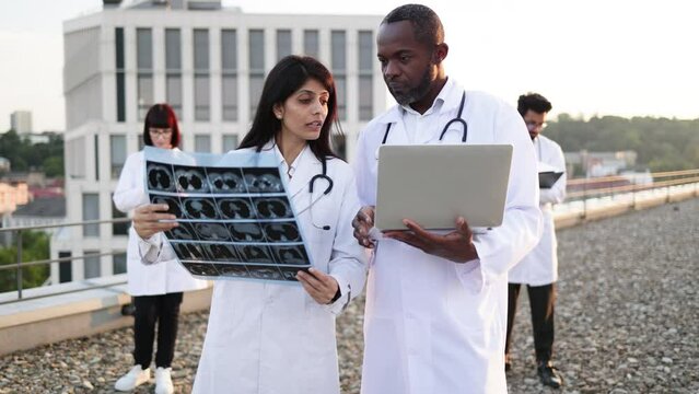 Portrait of bearded African male with laptop in lab coat and young Indian female doctor with stethoscope looking at camera and smiling on roof. Medical workers talking about patient examinations.