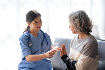 Asian nurse in blue scrubs gives a glass of water to an elderly woman with gray hair in a bright room.