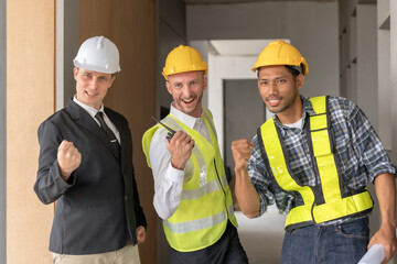 Three happy men in helmets and safety vests are at a work site, smiling and raising their fists up.