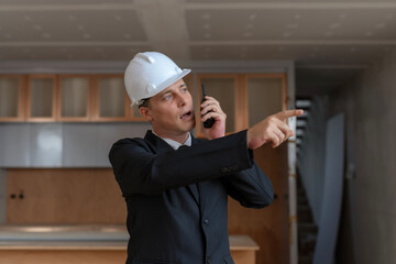 Professional architect in suit and helmet talks on a walkie-talkie, pointing directions on a construction site.