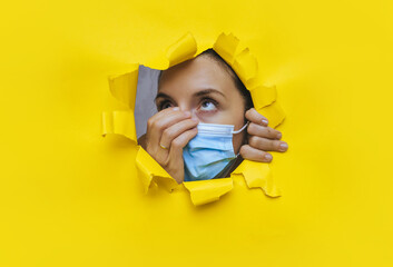 Surprised young woman looks through a torn hole in yellow paper and adjusts a medical mask. The...