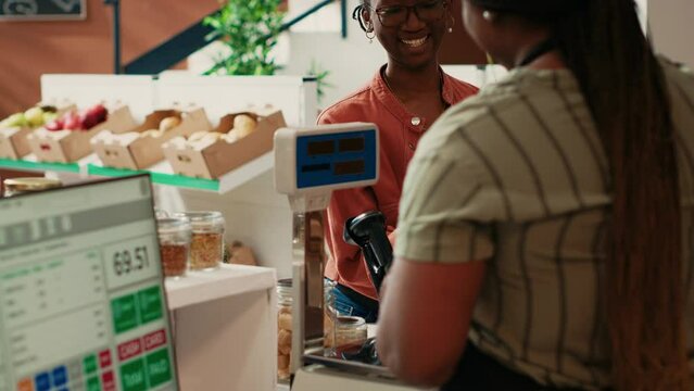 Organic store merchant scanning jars for customer at checkout, selling homemade additives free sauces and grains in recyclable jars. African american buyer looking for fresh natural pantry supplies.