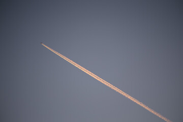 A jet plane in the blue sky. White contrail in sunset orange colors. Beautiful moment at sunset.